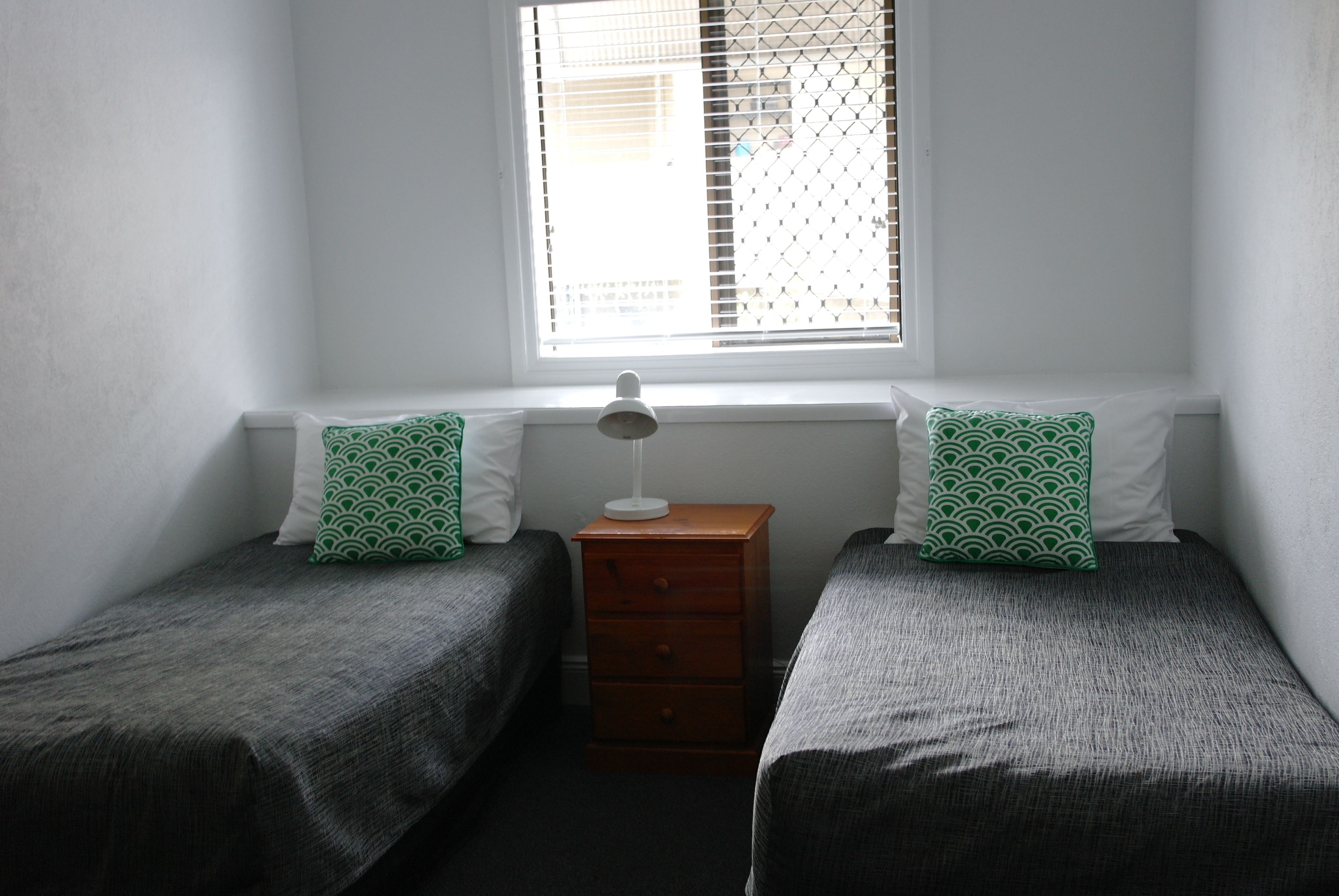 A small, simple room with two single beds, a nightstand, and a window with blinds.
