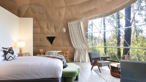 A spacious bedroom with a large bed, a green ottoman, a seating area with chairs, and a view of trees through a large window. - Jackson Ranch by Bannisters (Bawley Point)