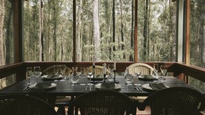 A dining table set for six with chairs, set against a forest backdrop through large windows. - Jackson Ranch by Bannisters (Bawley Point)