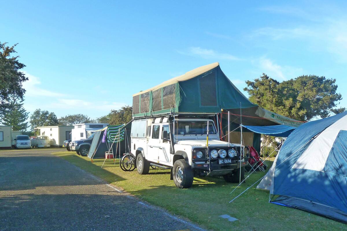 Een camping met een witte SUV geparkeerd in de buurt van een tent, een caravan en een bord met de tekst 'caravan park'.