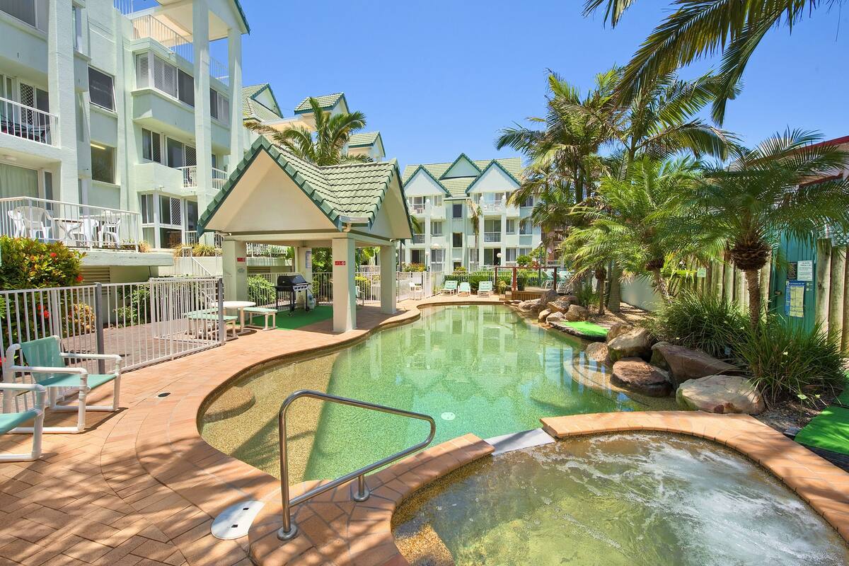 A pool area with lounge chairs and a small pavilion, surrounded by palm trees and a multi-story building.