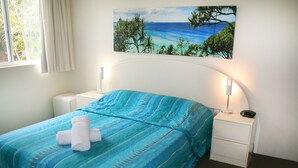 A neatly made bed with a blue bedspread and white towels, a painting of a tropical beach on the wall, and two bedside lamps on white nightstands. - The Bay Apartments (Coolangatta)