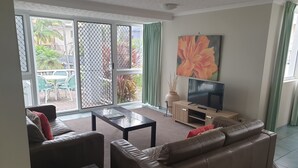 A living room with a sofa, coffee table, TV, and a view through a sliding glass door to an outdoor area with a table and chairs. - The Bay Apartments (Coolangatta)