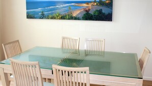 A dining area with a glass-top table and wooden chairs, a beach scene painting on the wall, and a view of the ocean. - The Bay Apartments (Coolangatta)