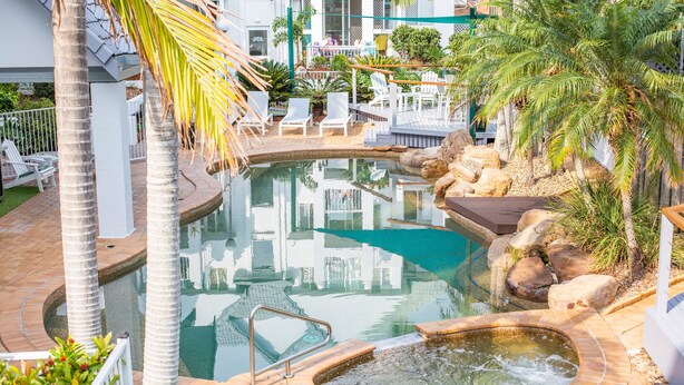 A swimming pool with a hot tub, surrounded by palm trees and lounge chairs. - The Bay Apartments (Coolangatta)