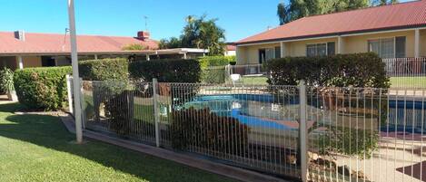 Un patio trasero cercado con piscina, un porche cubierto y una casa de techo rojo.