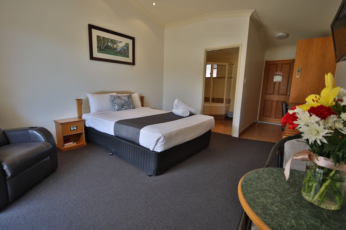 A hotel room with a bed, a nightstand, a chair, a small table with a vase of flowers, and a door leading to another room.