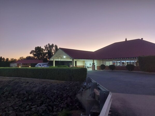 A building with a red roof and a covered entrance area. There are cars parked in front, and a pile of rocks is visible in the foreground.