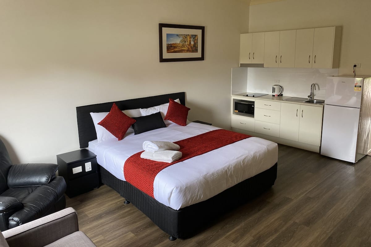 A hotel room with a bed, a nightstand, a chair, a kitchenette, and a framed picture on the wall.