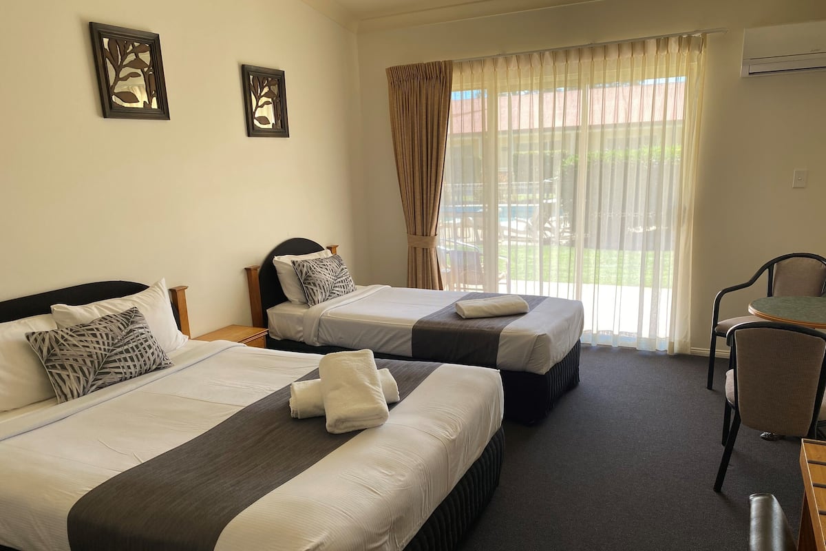 A hotel room with two beds, a desk, and a chair. There are two framed pictures on the wall, a window with curtains, and an air conditioning unit.