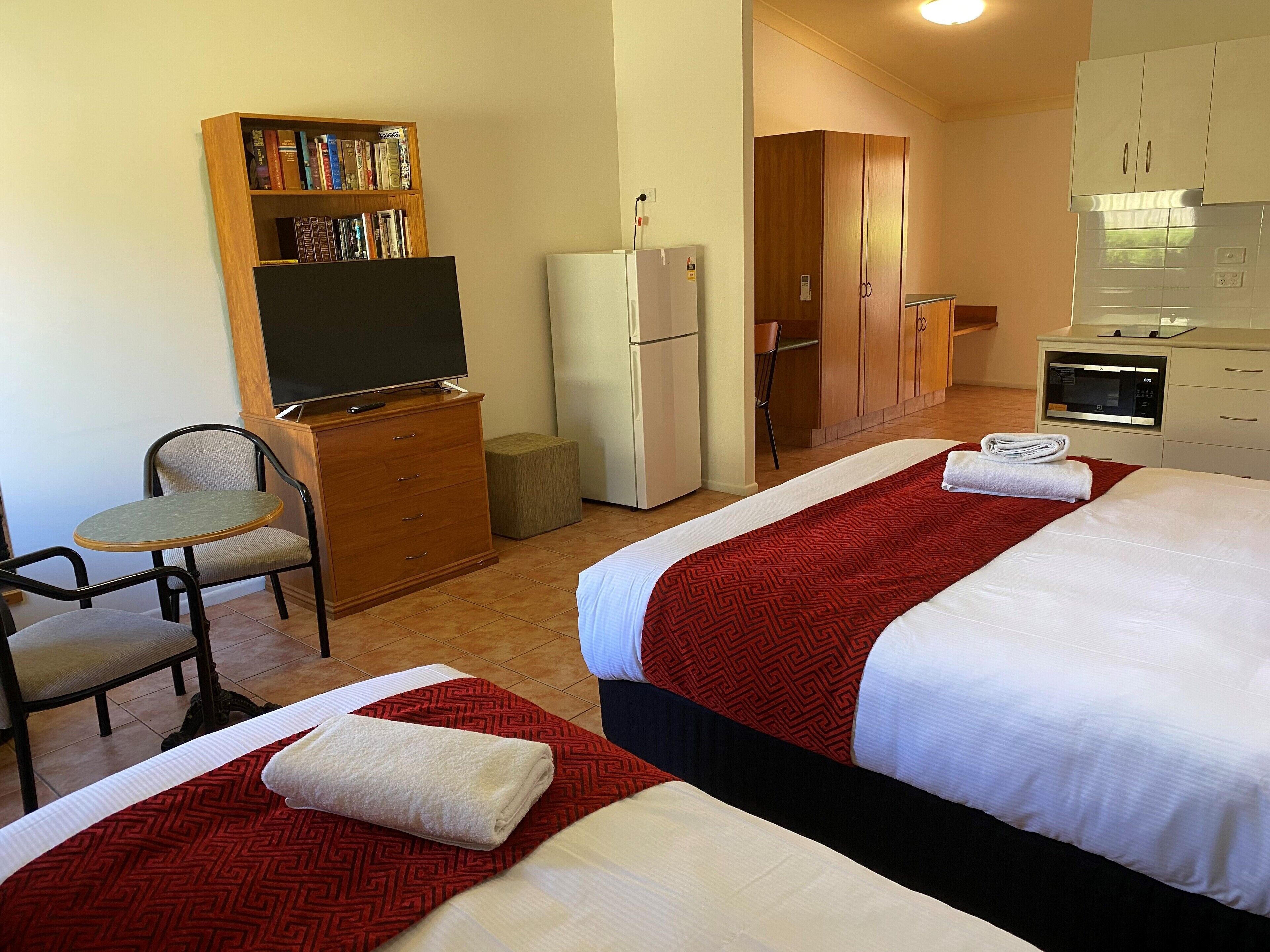 A hotel room with two beds, a small desk with a chair, a bookshelf, a refrigerator, a microwave, and a kitchenette.