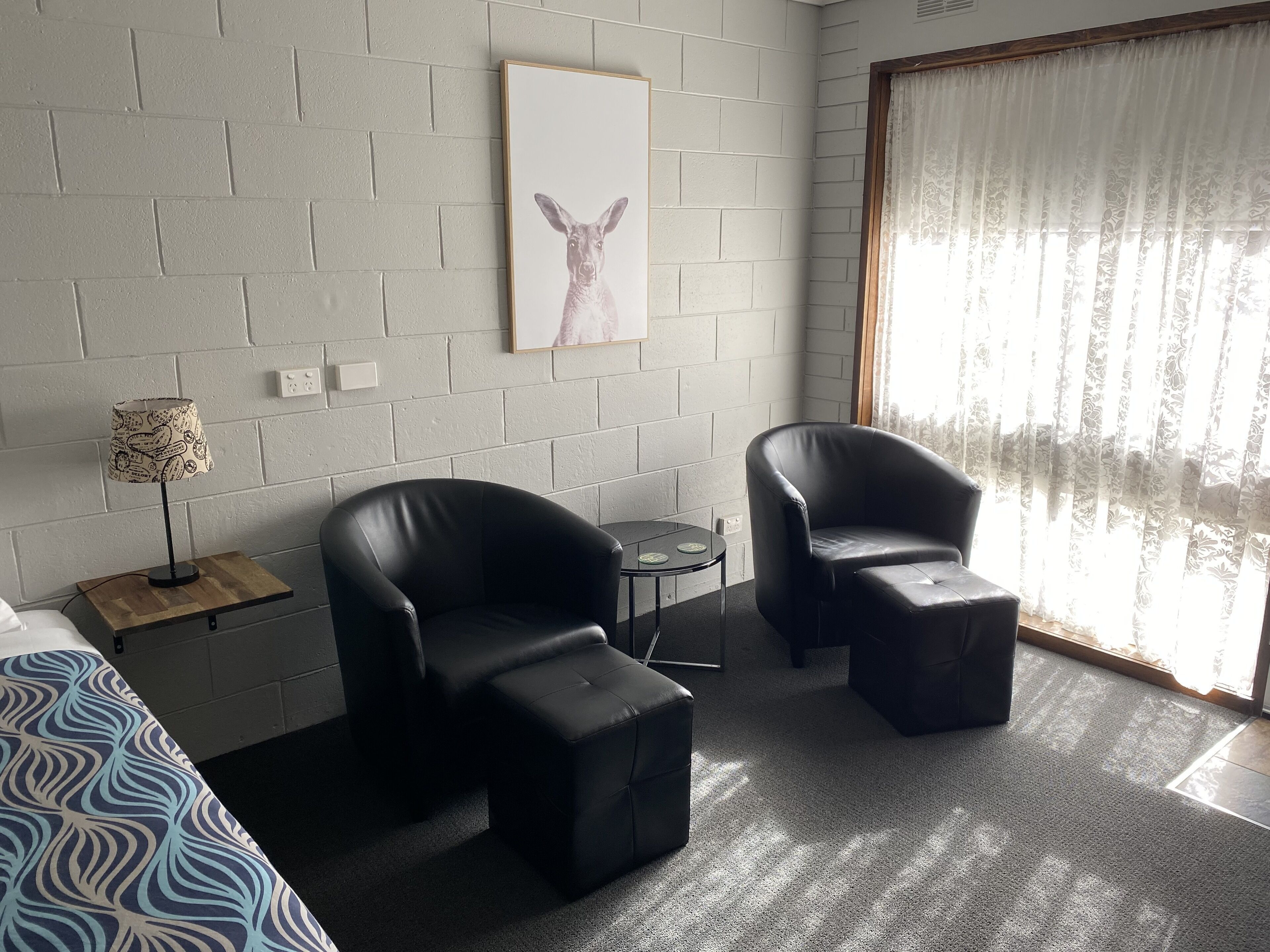 A hotel room with two black leather armchairs, a small round table, a lamp with a patterned lampshade, a framed picture on the wall, and a large window with sheer curtains.