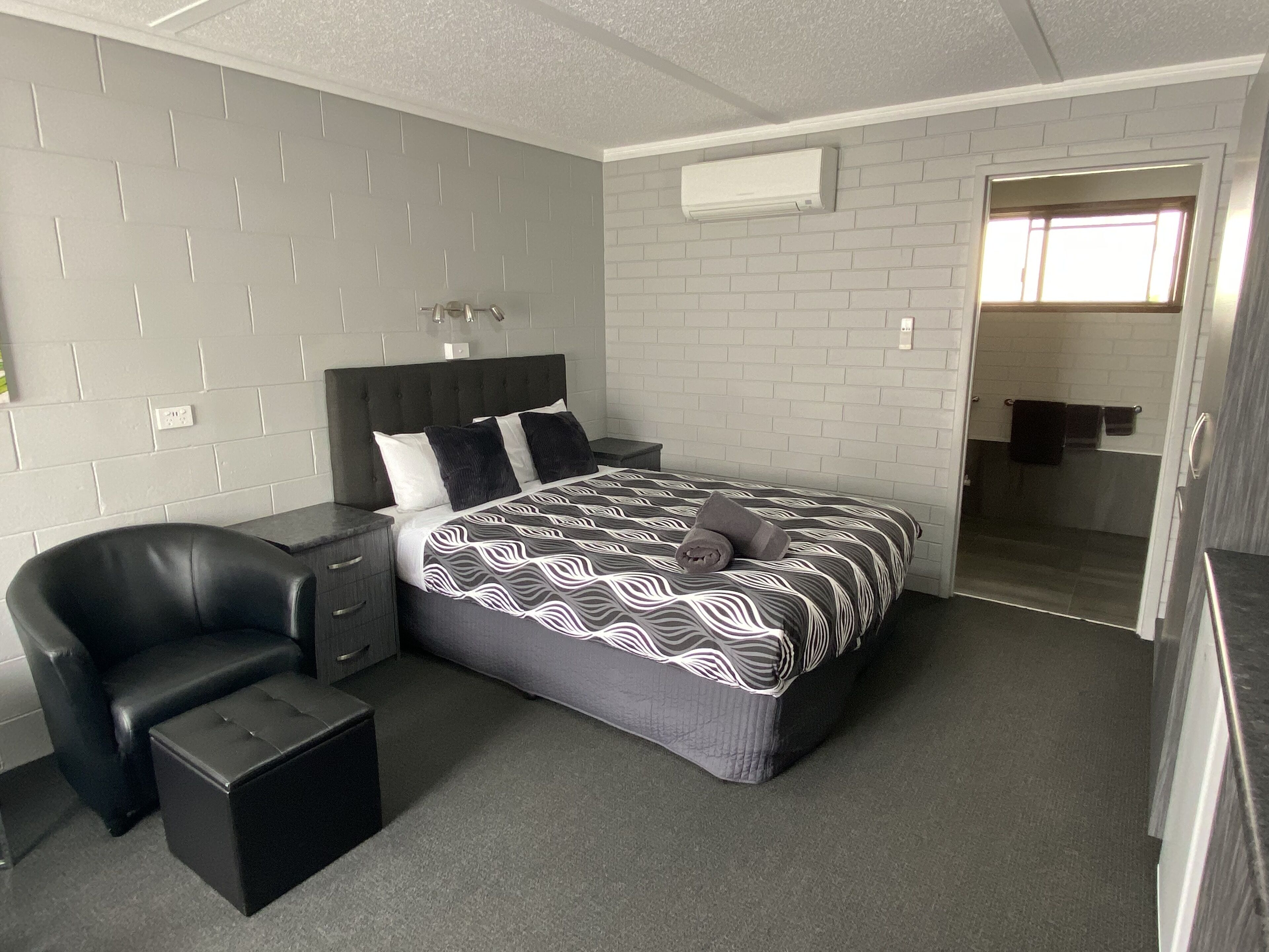 Una habitación de hotel moderna con pared de ladrillo gris, una cama grande con colcha estampada, un sillón de cuero negro, una mesita de noche pequeña y una ducha a ras de suelo visible a través de una puerta abierta.