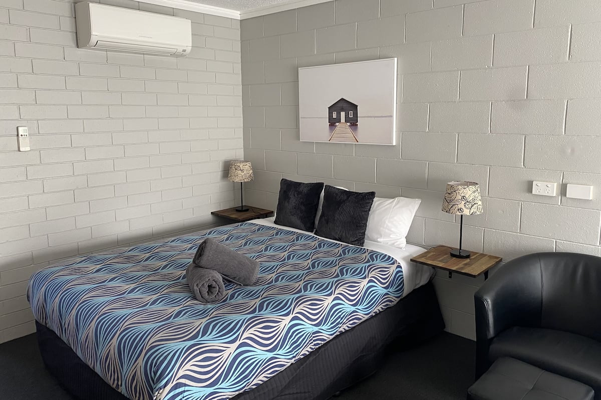 A bedroom with a bed, two nightstands, a chair, and a wall-mounted air conditioner.