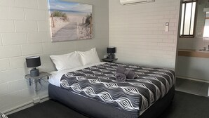 A modern hotel room with a large bed, a painting of a beach, a small bedside table with a lamp, and a bathroom visible through an open door. - Mariner Motel (Portland)