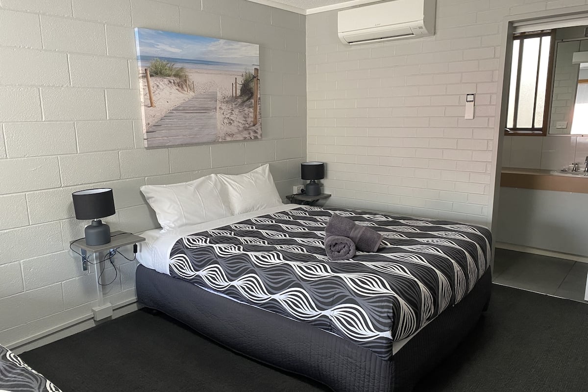 A modern hotel room with a large bed, a painting of a beach, a small bedside table with a lamp, and a bathroom visible through an open door.