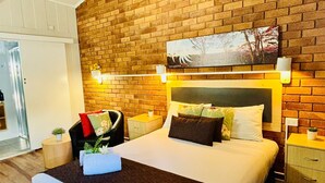 A modern hotel room with a brick wall, a large bed with multiple pillows, a nightstand, and a small table with a plant. - Downs Motel (South Toowoomba)
