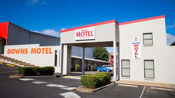 A motel with a sign reading "DOWNS MOTEL" and a phone number. There are parking spaces and a blue car parked in front. - Downs Motel (South Toowoomba)