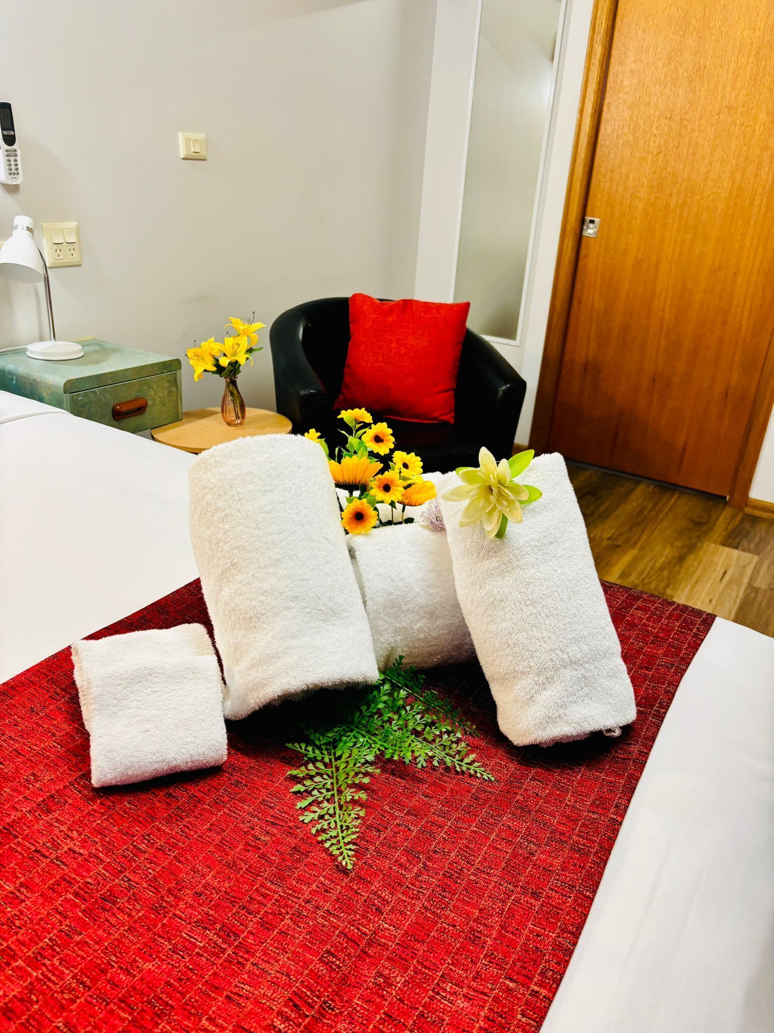 A hotel room with a bed, a red and white checkered area rug, a small table with a vase of yellow flowers, and a chair with a red cushion.