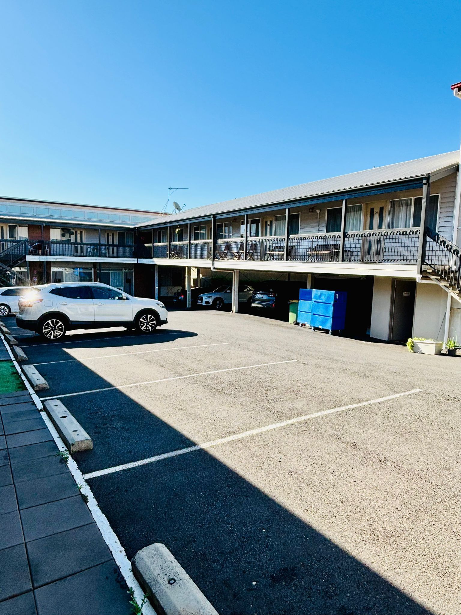 A two-story motel with a parking lot, a white car parked in the lot, and a blue dumpster.