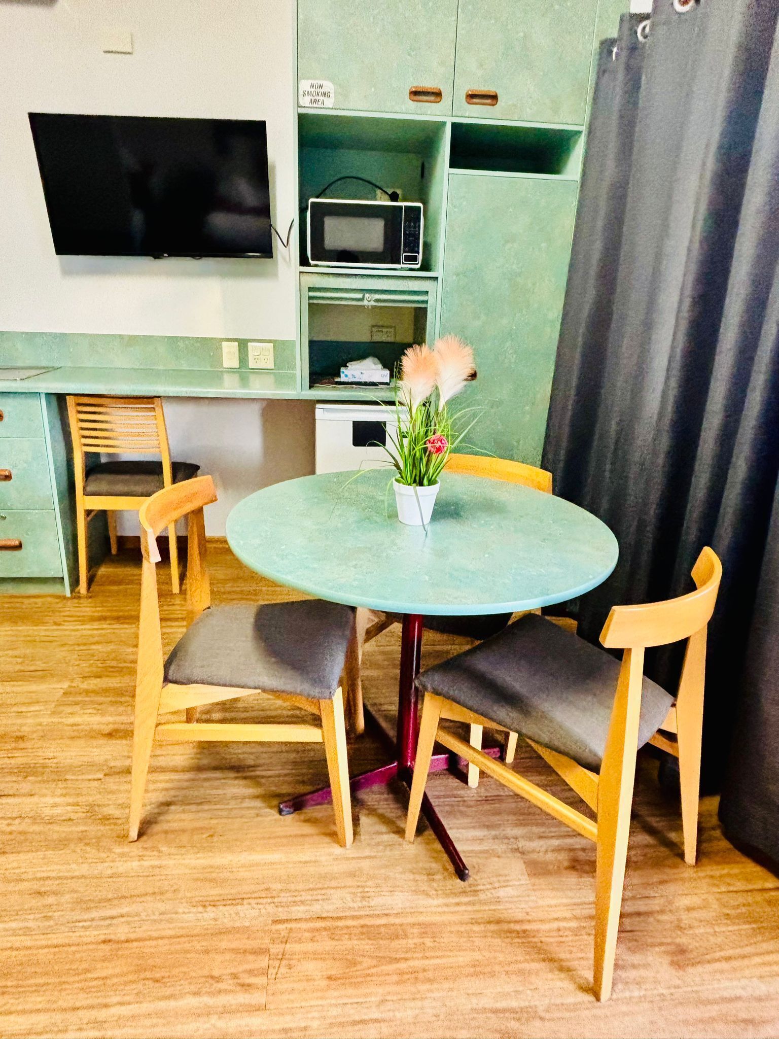 Une cuisine compacte équipée d’un four à micro-ondes, d’un petit réfrigérateur et d’un téléviseur fixé au mur. On y trouve une table ronde avec deux chaises, ainsi qu’une plante en pot.