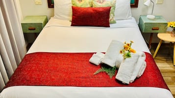 Un lit bien fait, avec une couverture rouge, des oreillers blancs et un coussin à motifs floraux. On y trouve deux tables de chevet avec des lampes, une petite table ronde sur laquelle est posé un vase de fleurs, et une valise verte.