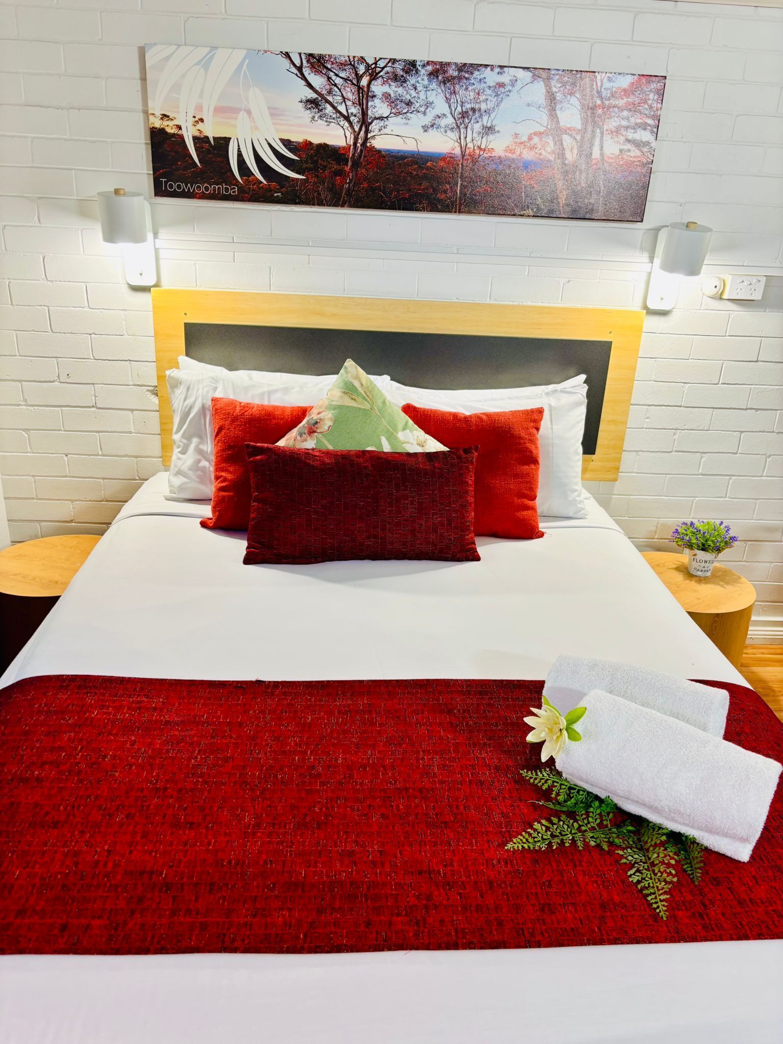 Ein Hotelzimmer mit Bett, roten und weißen Betttextilien, einem Holz-Kopfteil, einem Bild über dem Bett und einem kleinen Tisch mit einer Pflanze.