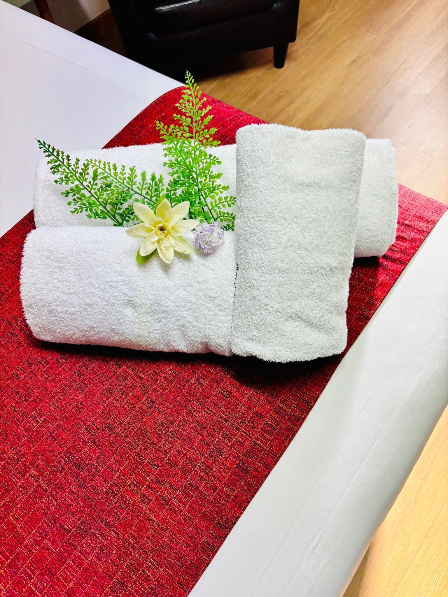 A neatly folded white towel with a decorative green leaf and a white flower, placed on a red textured surface.