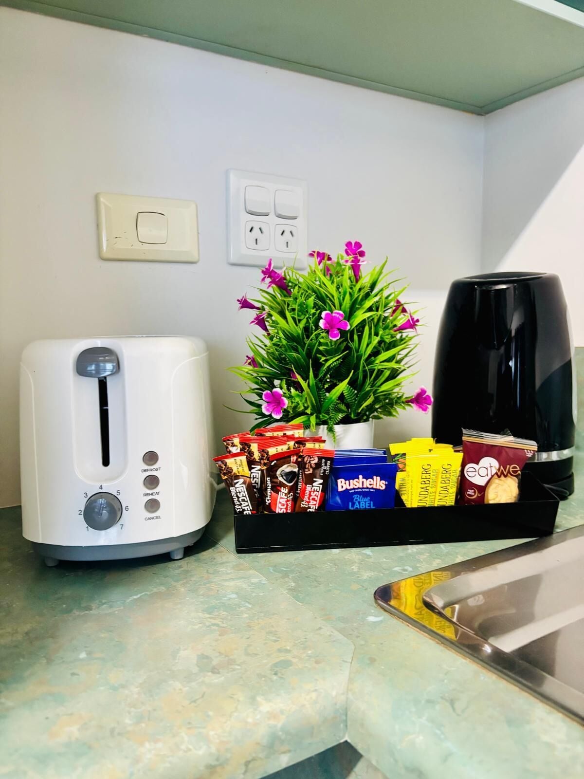 A kitchen counter with a white toaster, a black coffee maker, a potted plant with pink flowers, and a tray of coffee pods.