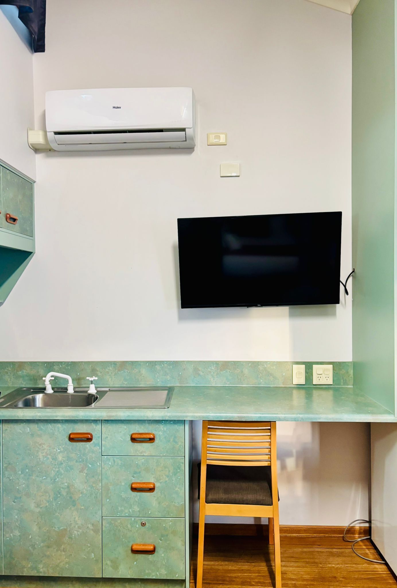 A compact kitchen with a built-in air conditioner, a flat-screen TV, a sink, and a wooden chair.