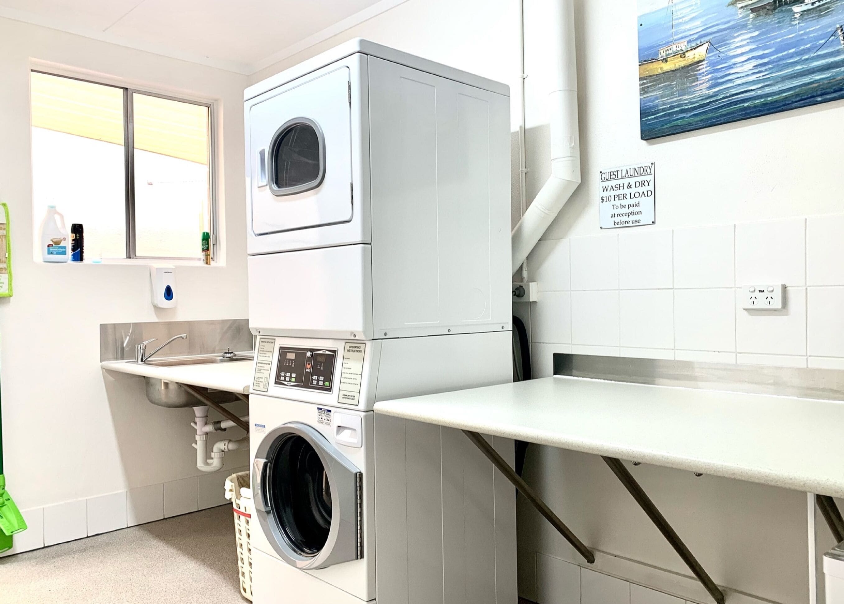 Une buanderie équipée de machines à laver et de sèche-linge superposés, d’un évier et d’un tableau représentant un bateau accroché au mur.