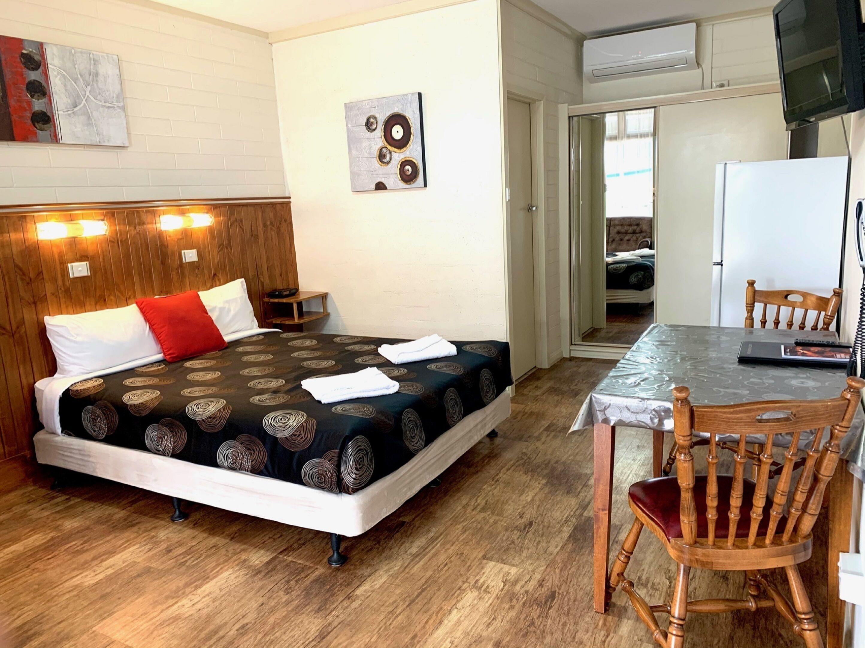Habitación de hotel con cama, cabecera de madera, una mesita con silla y una cocina visible a través de una puerta abierta.