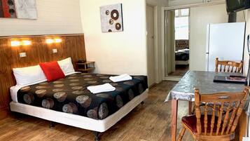 Habitación de hotel con cama, cabecera de madera, una mesita con silla y una cocina visible a través de una puerta abierta.