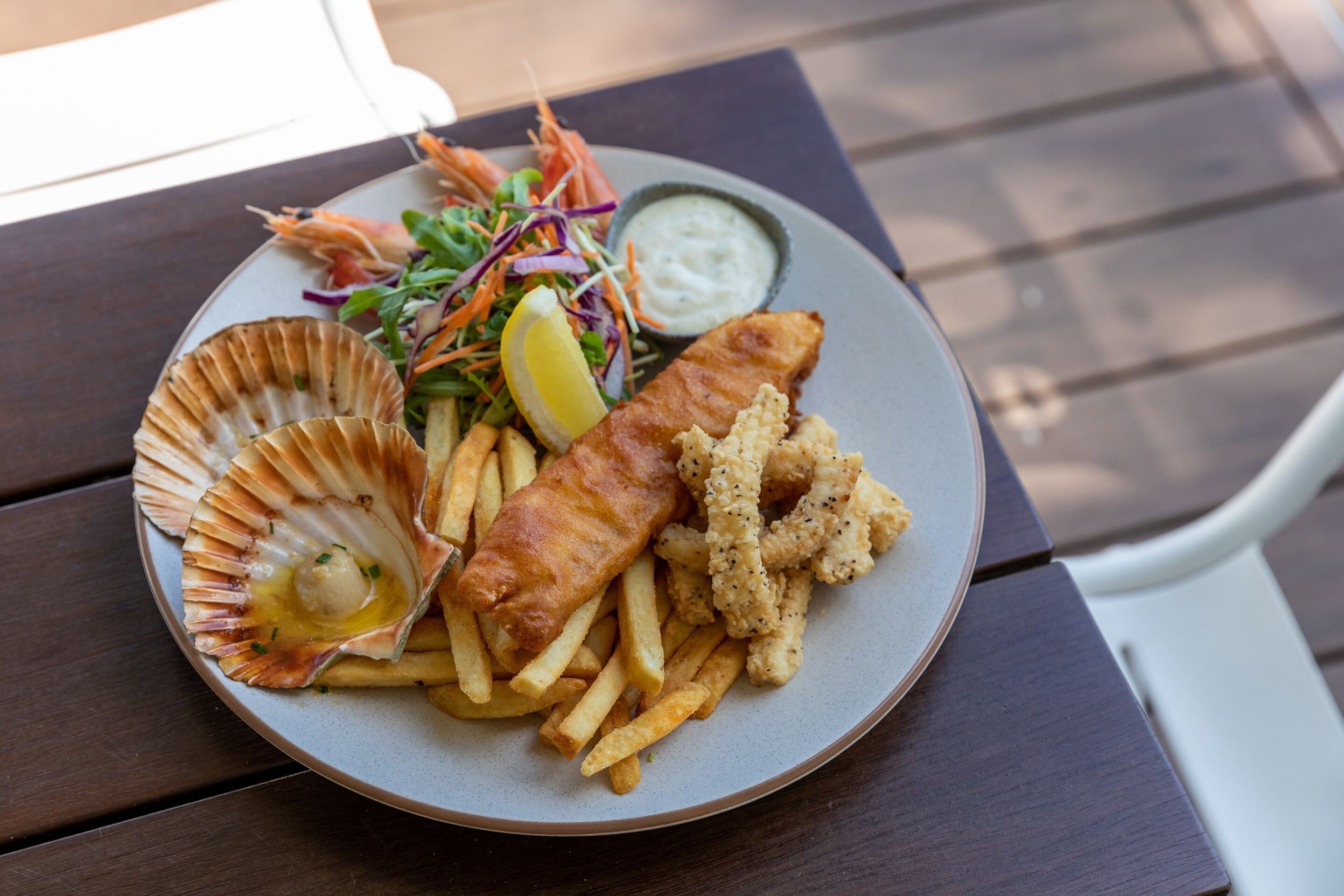 Ein Teller Fish and Chips mit einem gemischten Salat, einer Zitronenscheibe und einer kleinen Schüssel Sauce auf einem Holztisch.