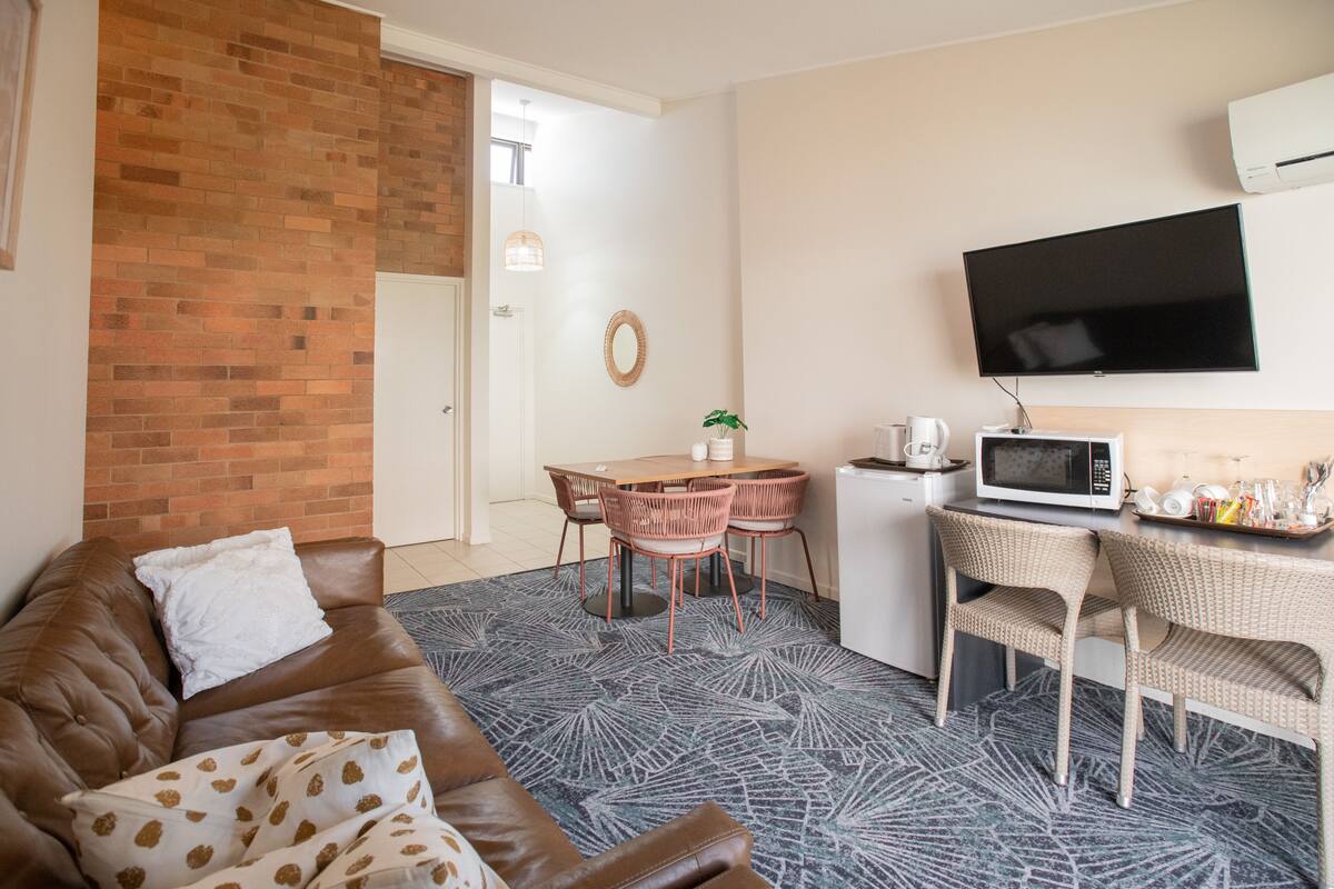 Una sala de estar con un sofá de cuero marrón, una mesa de comedor con sillas, un microondas y un televisor montado en la pared.