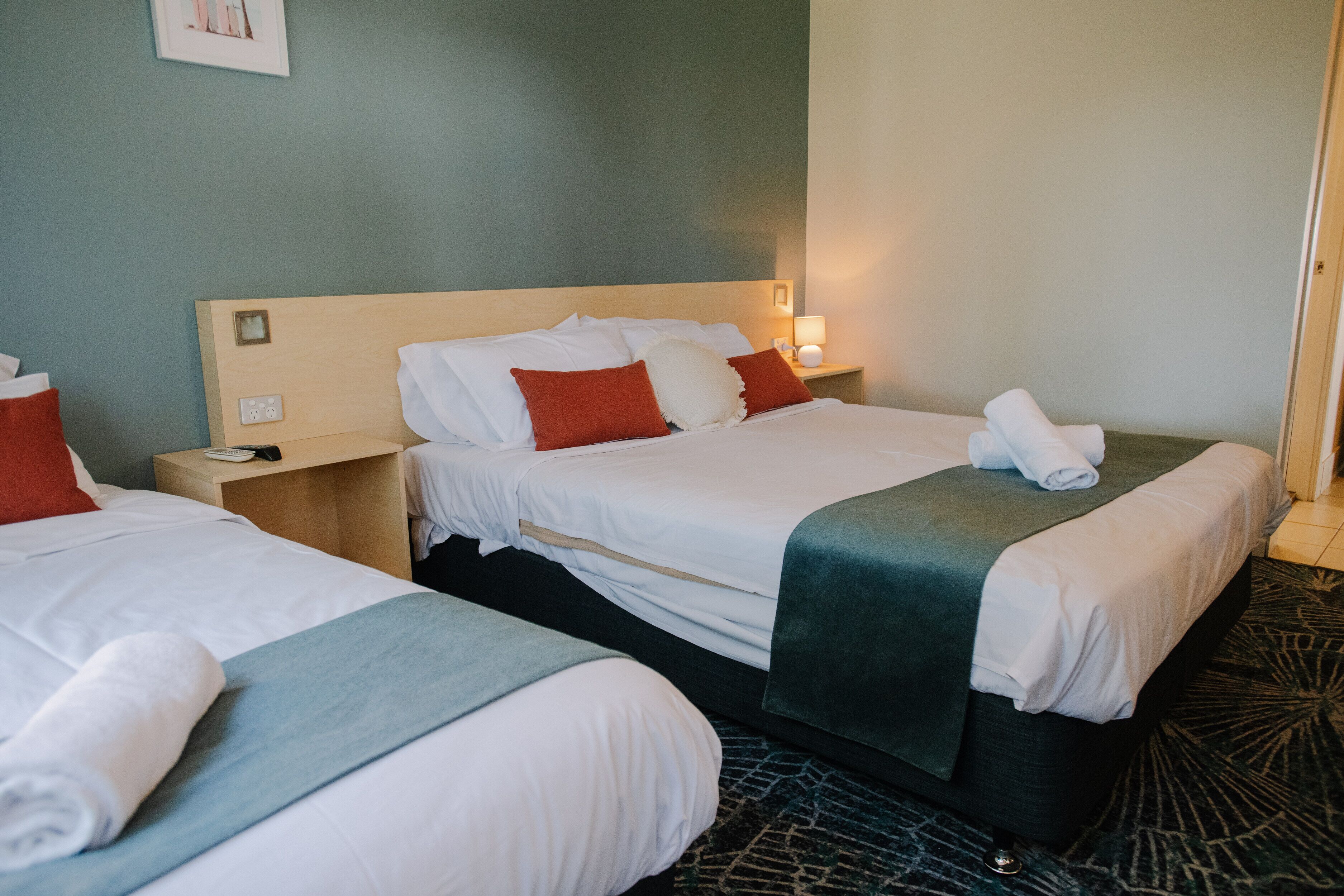 A hotel room with two beds, each with white linens, red and white pillows, and a green throw blanket. There is a nightstand with a lamp and a phone.