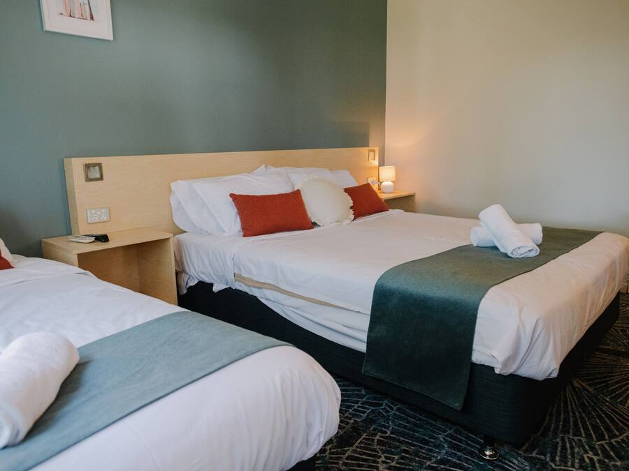 A hotel room with two beds, each with white linens, red and white pillows, and a green throw blanket. There is a nightstand with a lamp and a phone.