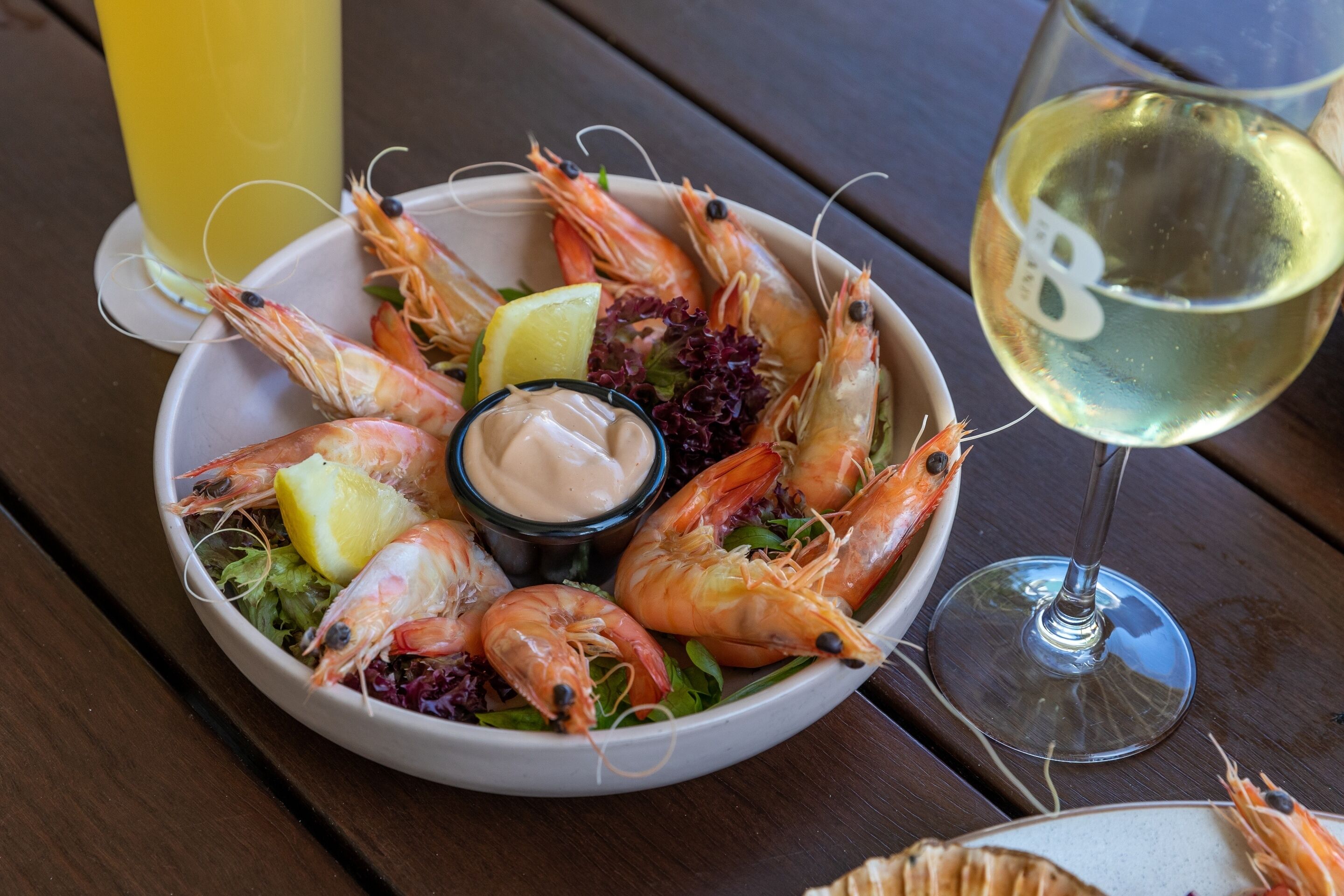 A plate of cooked shrimp with lemon slices, a small bowl of sauce, and a glass of white wine on a wooden table.
