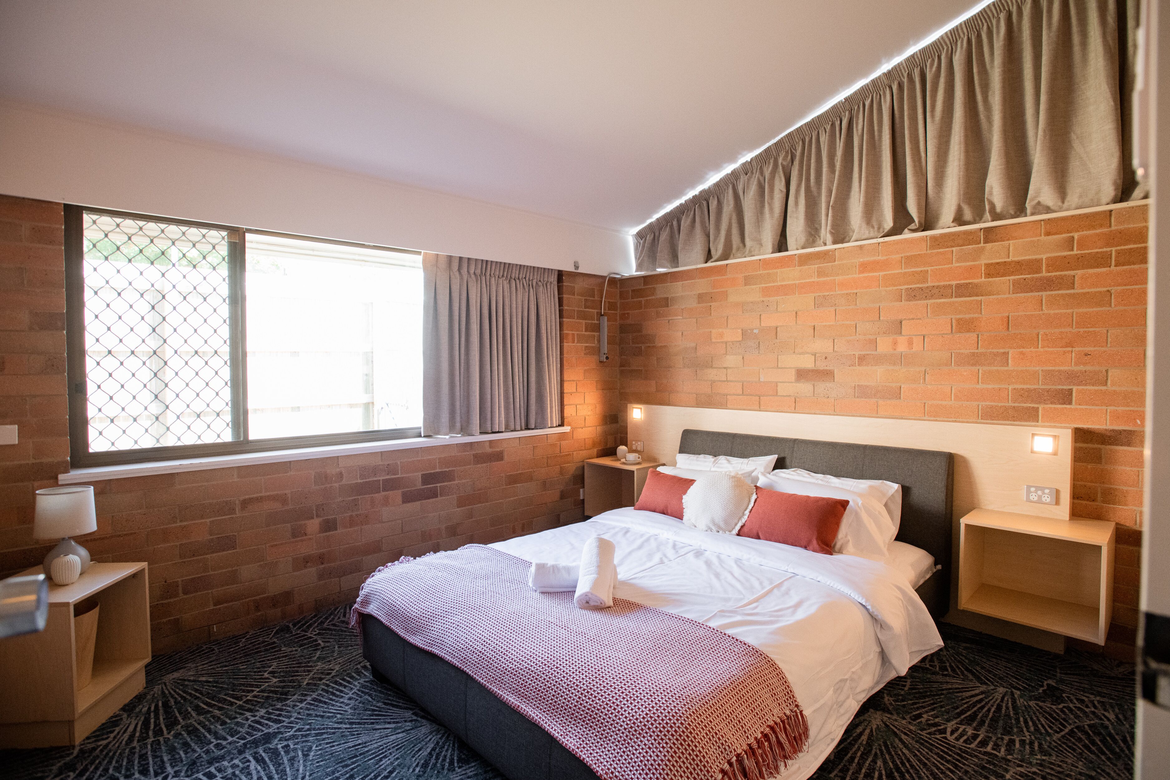 A hotel room with a brick wall, a large window with curtains, a neatly made bed, and a small bedside table with a lamp.