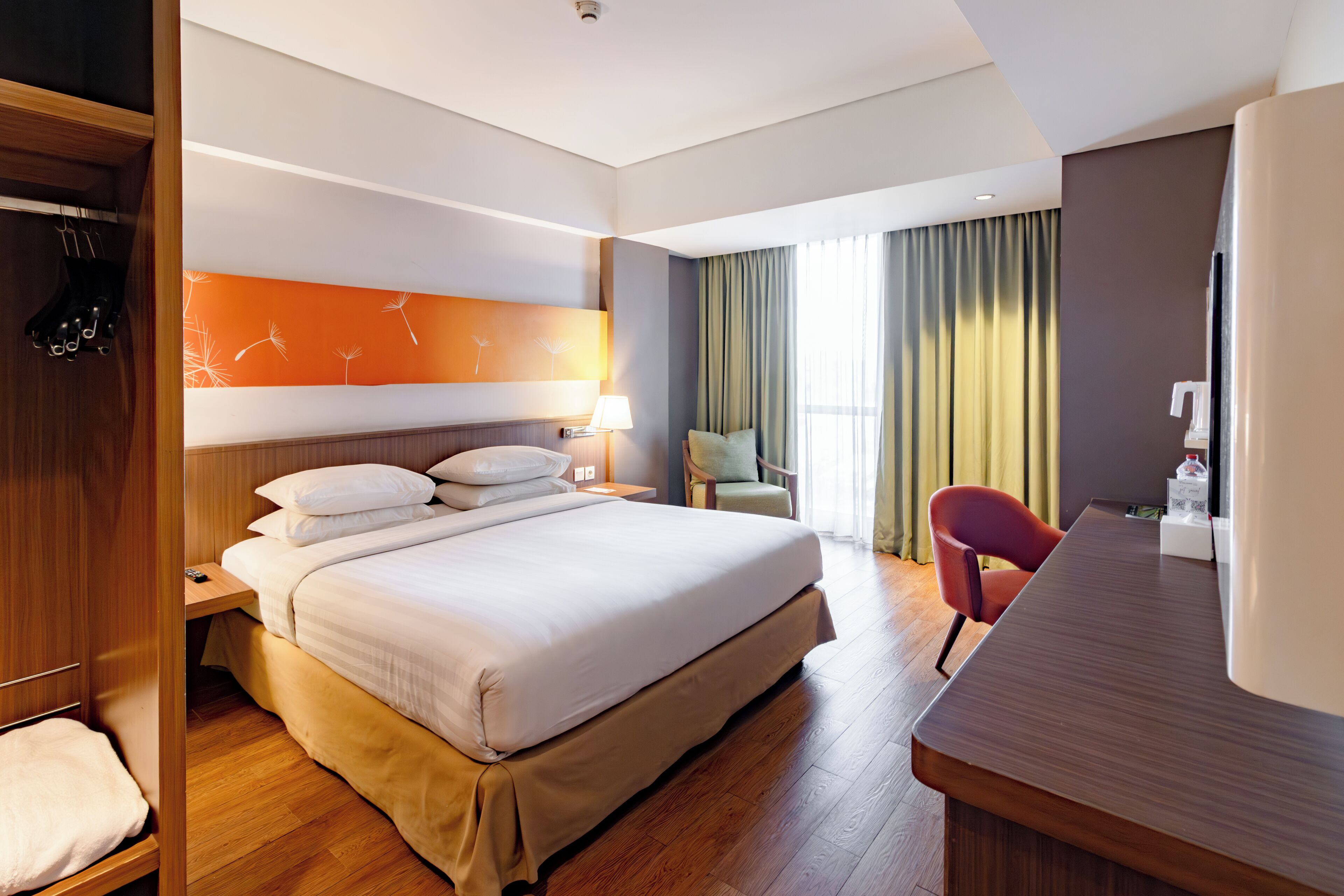 A hotel room with a large bed, wooden floor, a desk with a chair, a wardrobe, and a view of the outdoors through curtains.