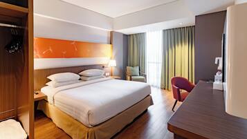 A hotel room with a large bed, wooden floor, a desk with a chair, a wardrobe, and a view of the outdoors through curtains.
