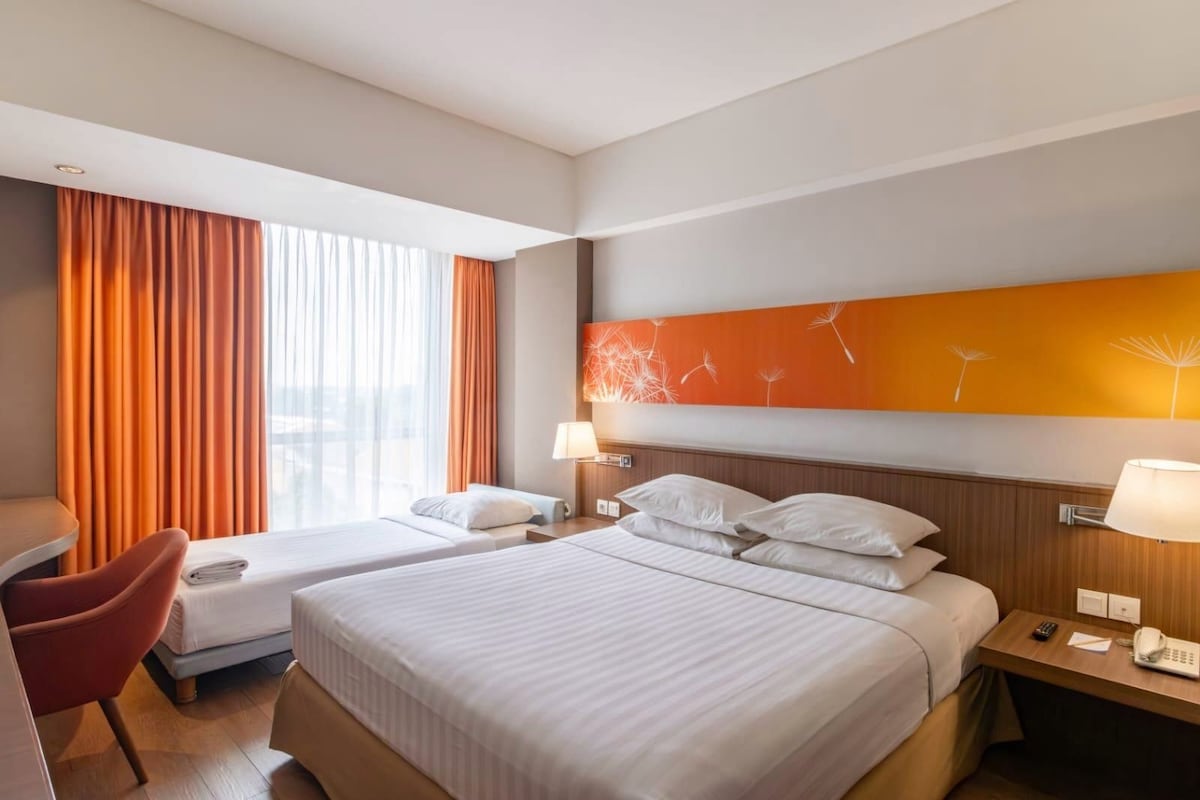 Habitación de hotel con dos camas, un escritorio, una silla y un ventanal con cortinas.