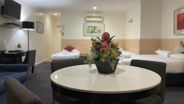 Una camera d'albergo con un tavolo rotondo, due poltrone, un letto con biancheria bianca, una TV a muro e un vaso di fiori sul tavolo.