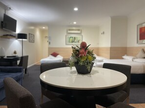 A hotel room with a round dining table, two armchairs, a bed with white bedding, a TV mounted on the wall, and a floral arrangement on the table. - Lakeview Motor Inn (Belmont South)