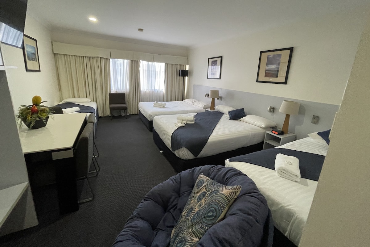 A hotel room with two beds, a desk, a chair, and a small table with a plant.