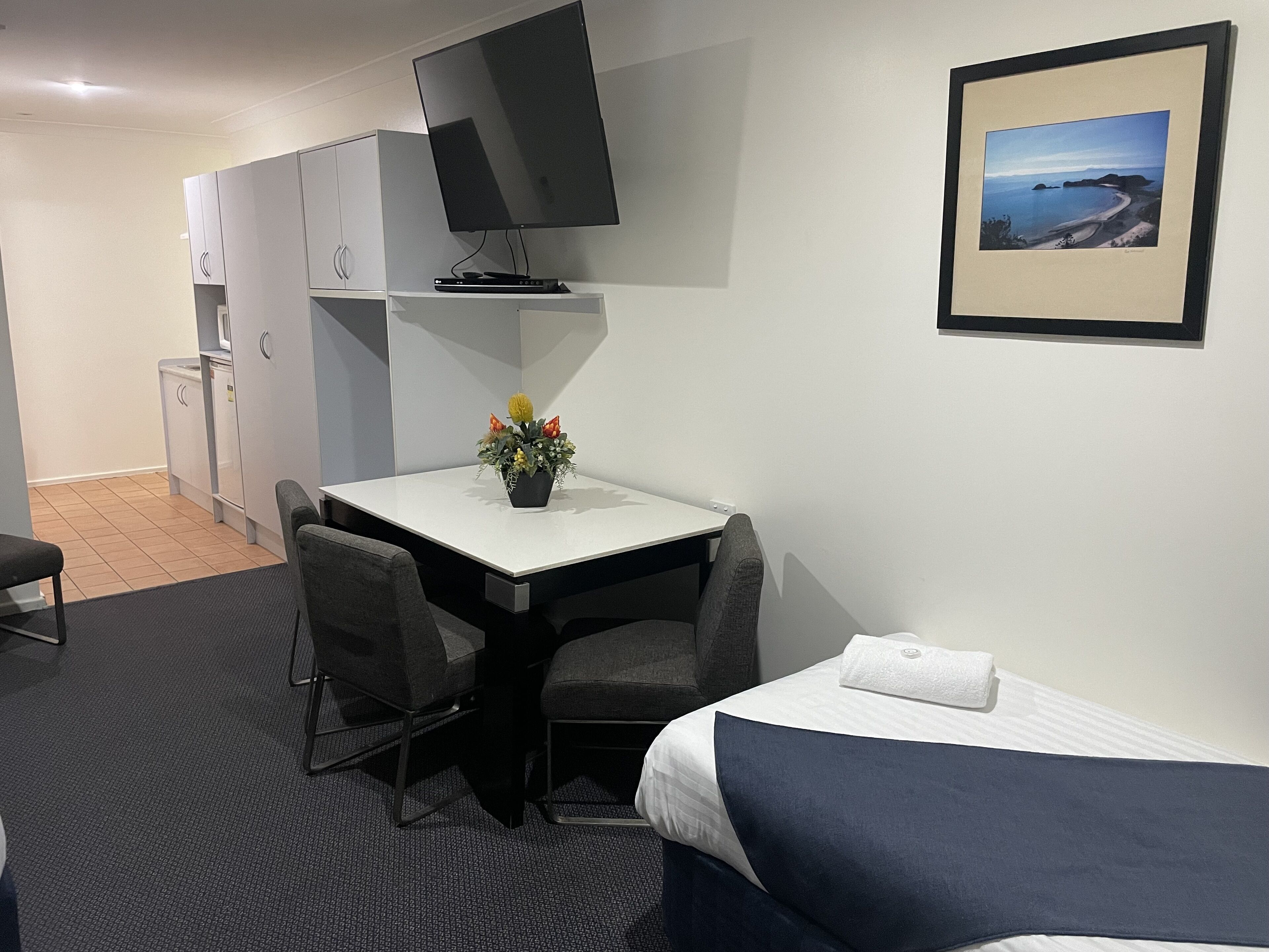 A hotel room with a bed, a desk with a chair, a television, a small table with a flower arrangement, and a framed picture on the wall.