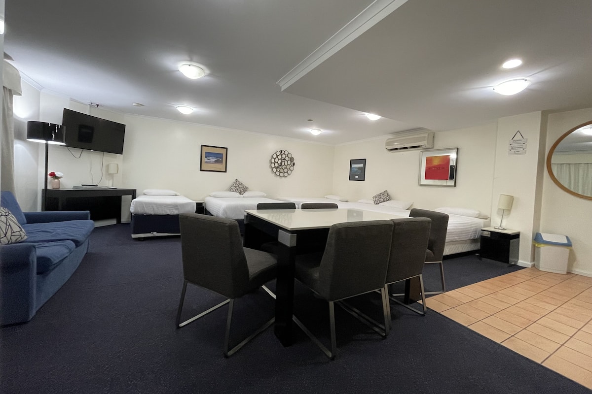 A hotel room with a dining table, four chairs, a sofa, a TV, and two beds.