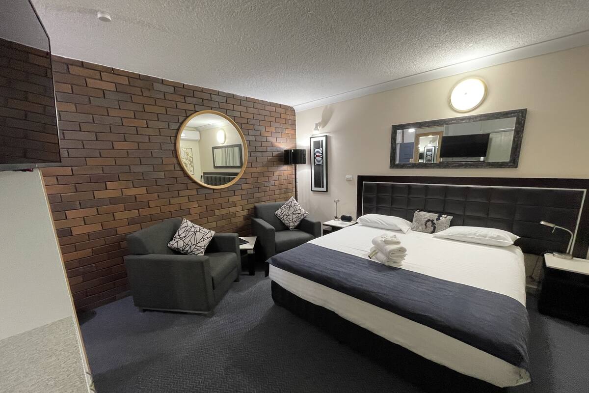 A hotel room with a bed, two armchairs, a small table, a mirror, and a brick wall.