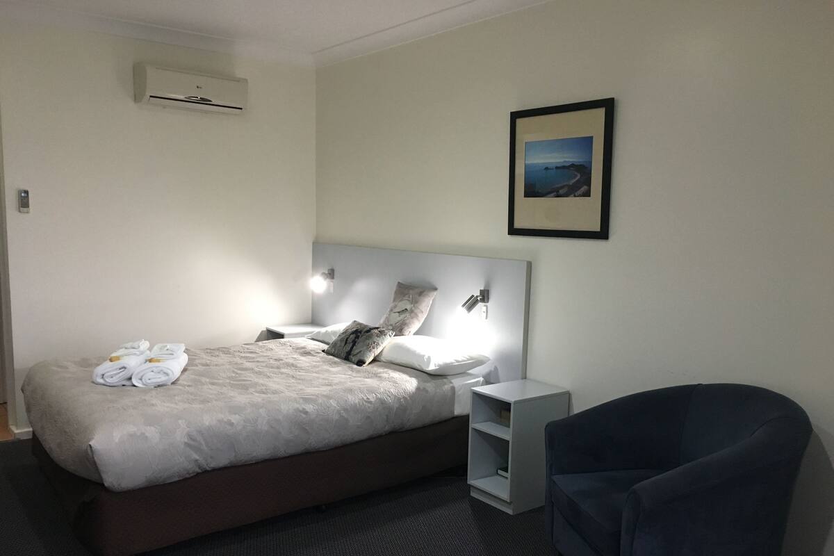 A hotel room with a bed, a chair, a nightstand, and a framed picture on the wall.