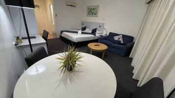 A hotel room with a bed, a sofa, a small round table, a desk, and a potted plant.